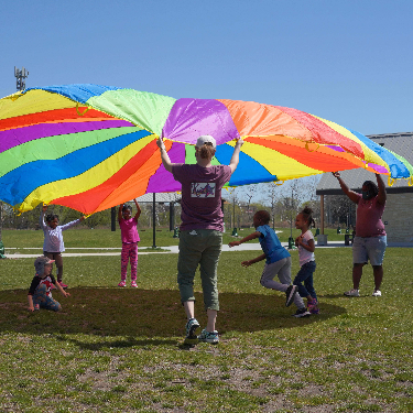 Kids running across grass underneath a lifted rainbow parachute